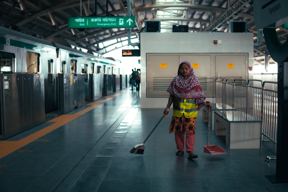 Two emergency cleaners from Cleaner Battersea are seen on a city street at Clapham Junction, dressed in high-visibility orange and red uniforms. They are pushing cleaning equipment, including a vacuum