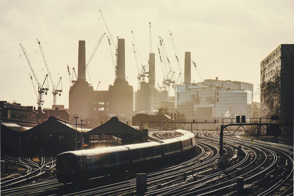 A view of the historic Battersea Power Station across the River Thames, featuring its distinctive red-brick exterior and four tall chimneys under a cloudy sky. Adjacent modern glass buildings reflect 