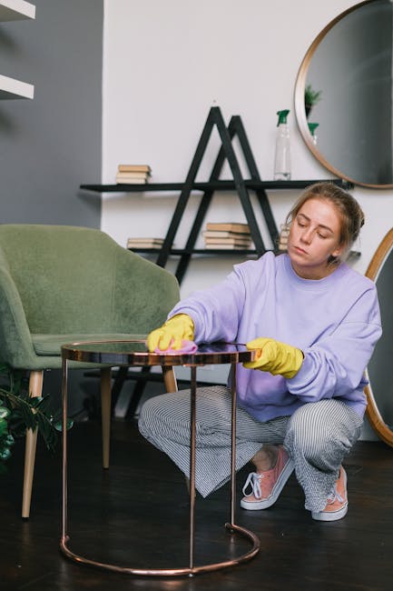 A young woman with light brown hair tied back, dressed in a lavender sweatshirt, patterned grey and white trousers, and yellow cleaning gloves, is kneeling on a dark wooden floor while wiping a small,